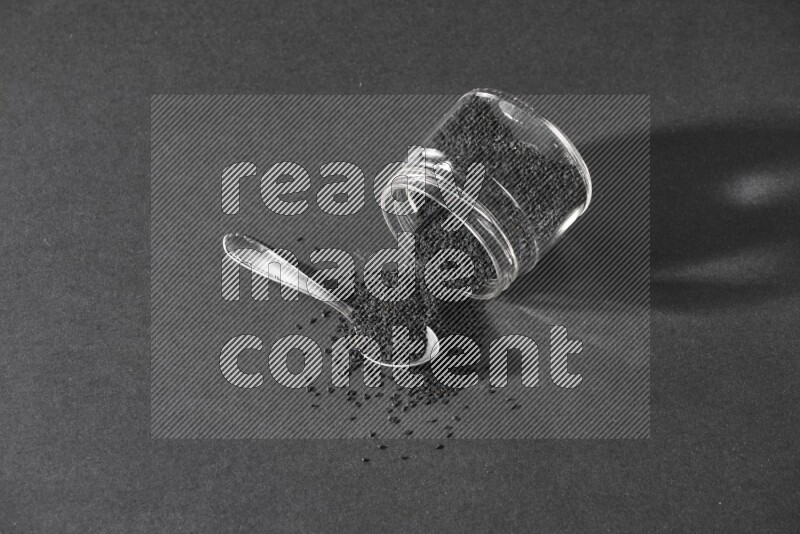 A flipped glass jar full of black seeds and seeds spread out with a metal spoon full of the seeds on a black flooring