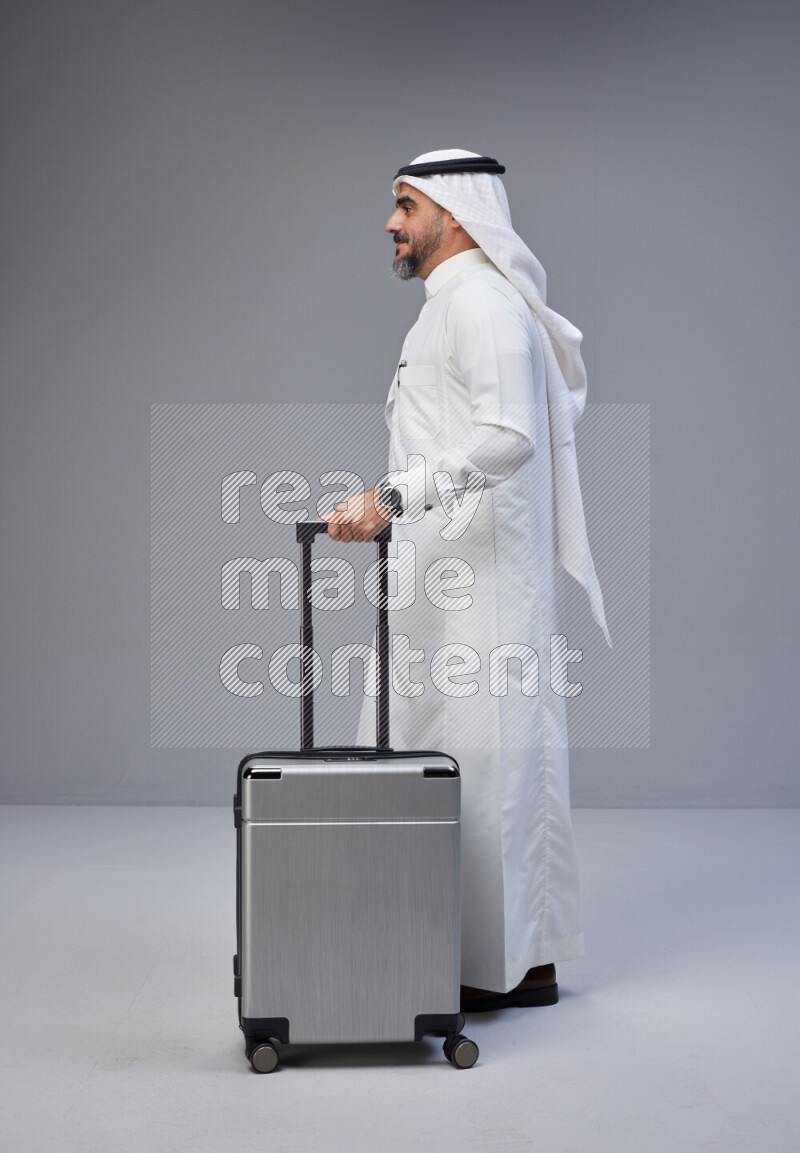 Saudi man wearing Thob and white Shomag standing holding Travel bag on Gray background
