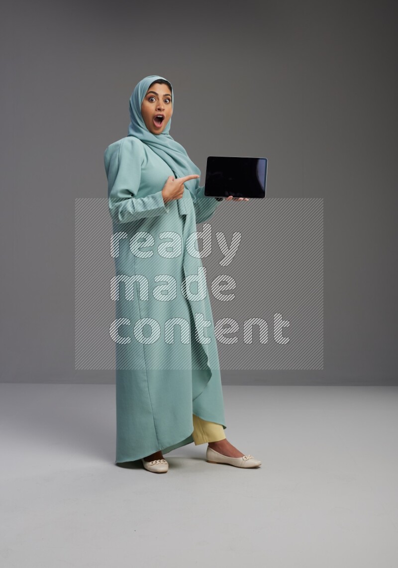 Saudi Woman wearing Abaya standing showing tablet to camera on Gray background