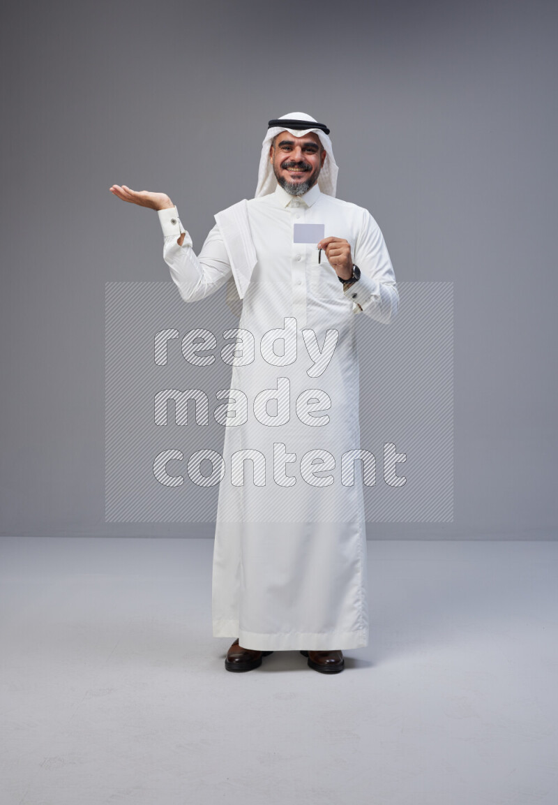 Saudi man Wearing Thob and white Shomag standing holding ATM card on Gray background