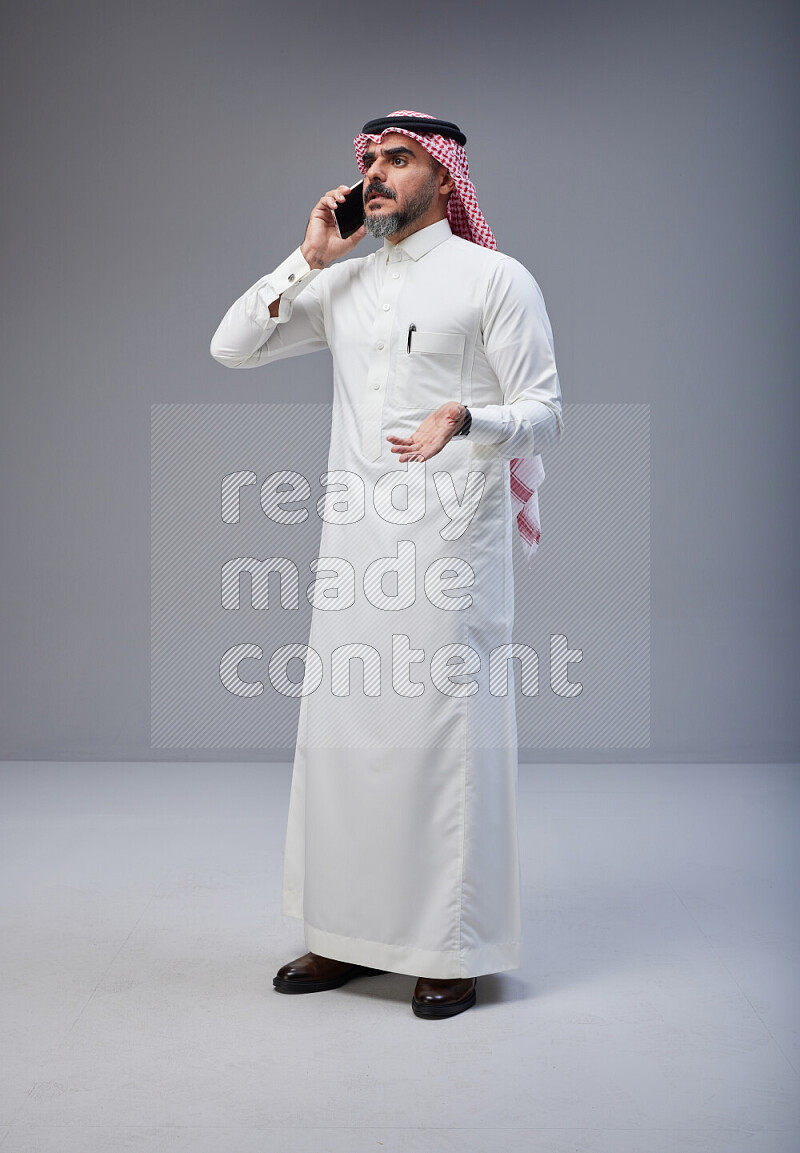Saudi man Wearing Thob and red Shomag standing talking on phone on Gray background