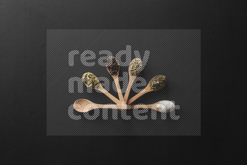 wooden spoons filled with white peppers, cloves, cardamom, salt, black peppers and basil on black flooring