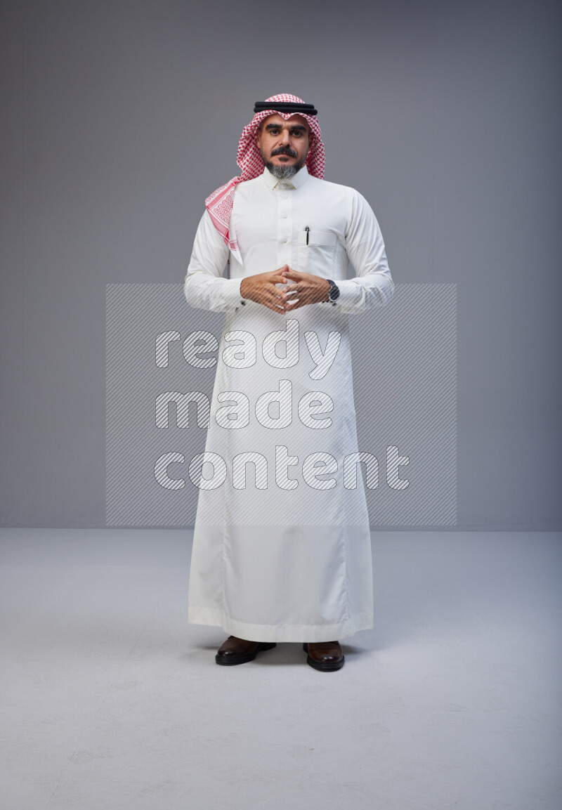 Saudi man Wearing Thob and red white Shomag standing interacting with the camera on Gray background