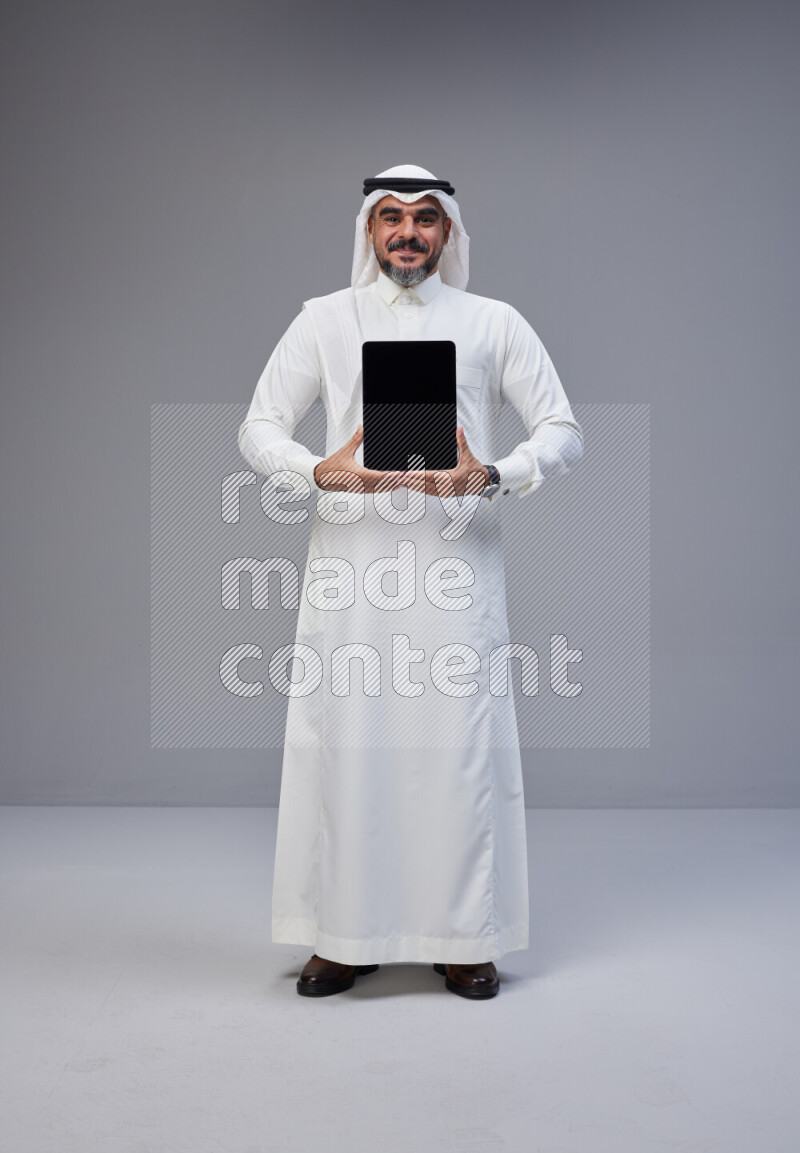 Saudi man Wearing Thob and white Shomag standing showing tablet to camera on Gray background