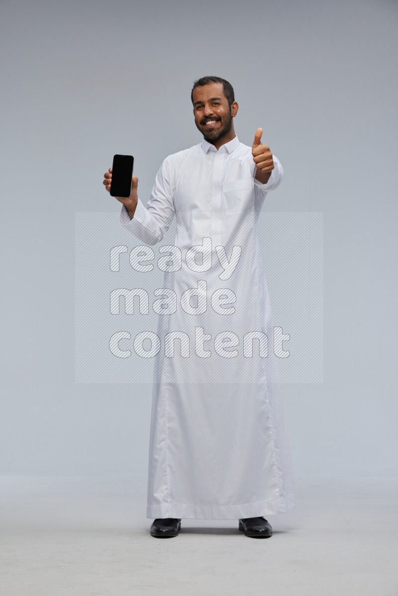 Saudi man wearing Thob standing showing phone to camera on Gray background