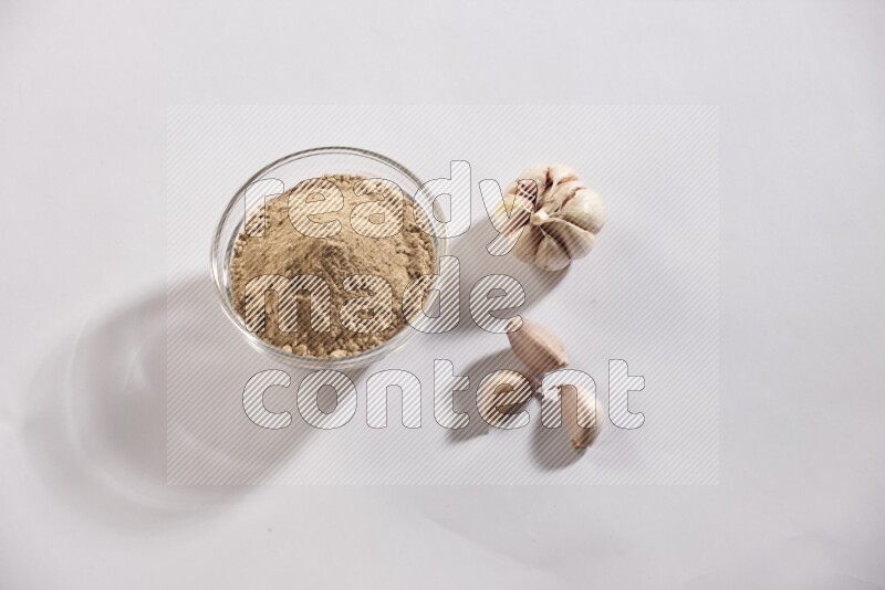 A glass bowl full of garlic powder with garlic bulb and some cloves beside it on a white flooring
