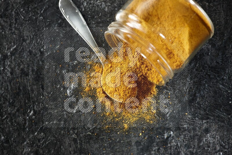 A flipped glass jar and a metal spoon full of turmeric powder and powder spilled out of it on textured black flooring