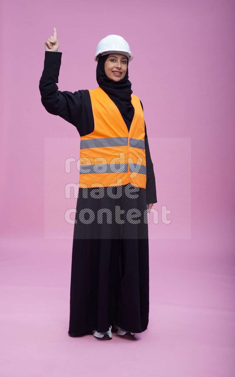 Saudi woman wearing Abaya with engineer vest and helmet standing interacting with the camera on pink background
