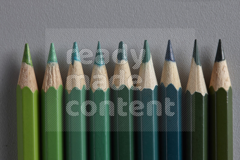 A collection of colored pencils arranged showcasing a gradient of green hues on grey background