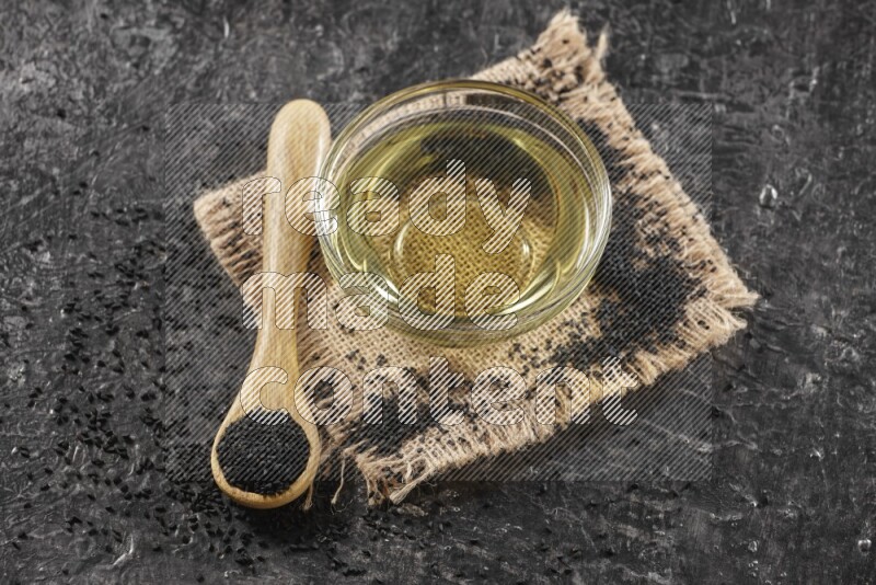A glass bowl full of black seeds oil and wooden spoon full of black seeds with seeds spreaded on burlap fabric on a textured black flooring