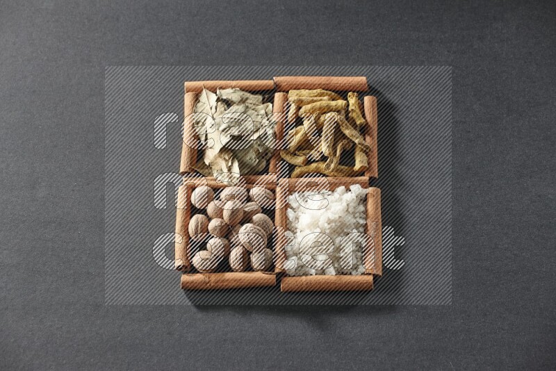 4 مربعات من عيدان القرفة ممتلئة بأوراق الغار والملح الأبيض وأصابع الكركم وجوزة الطيب على أرضية سوداء