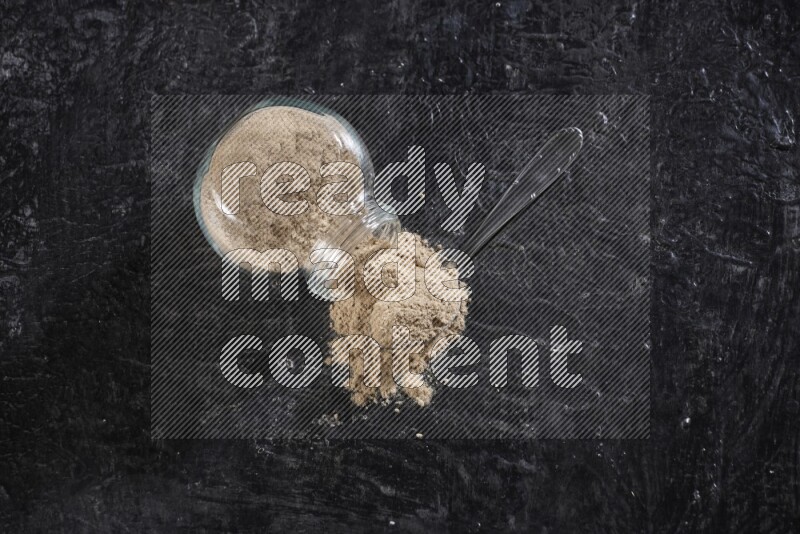 A glass spice jar full of garlic powder flipped and the powder came out with a filled metal spoon beside it on a textured black flooring