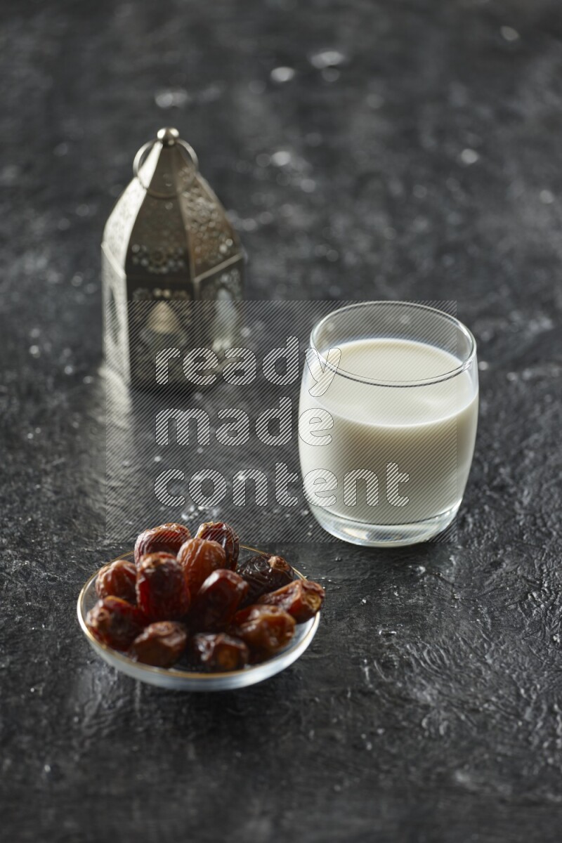 A silver lantern with different drinks, dates, nuts, prayer beads and quran on textured black background