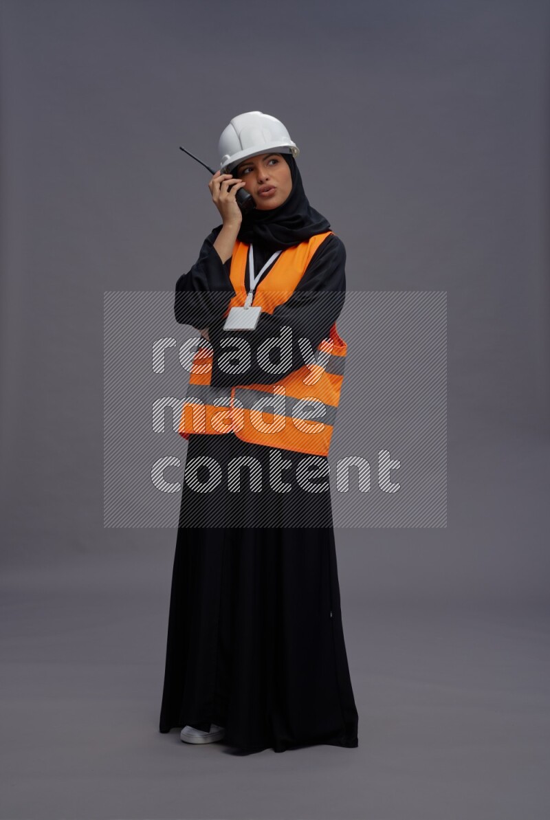 Saudi woman wearing Abaya with engineer vest with neck strap employee badge standing holding walkie-talkie on gray background