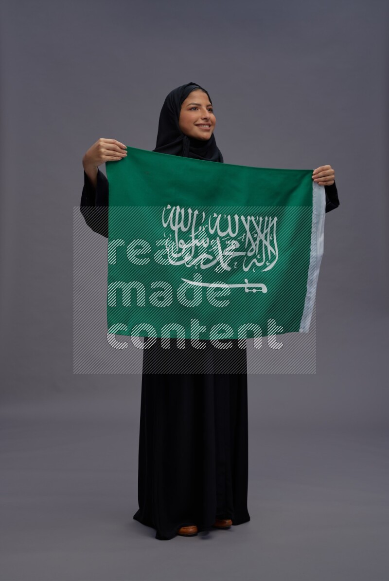 Saudi woman wearing Abaya standing holding Saudi flag on gray background