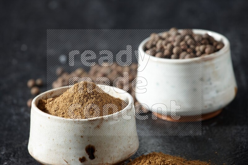 2 beige bowls, one full of allspice powder and the other full of whole balls and both spread on a textured black flooring