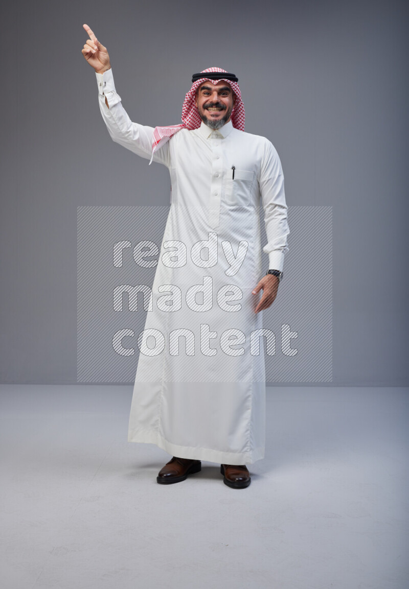 Saudi man Wearing Thob and red white Shomag standing interacting with the camera on Gray background