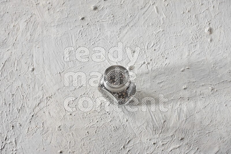 A glass spice jar full of black pepper on textured white flooring