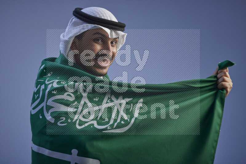 A close-up shot of Saudi man wearing thob and white shomag wrapping big Saudi flag on gray background