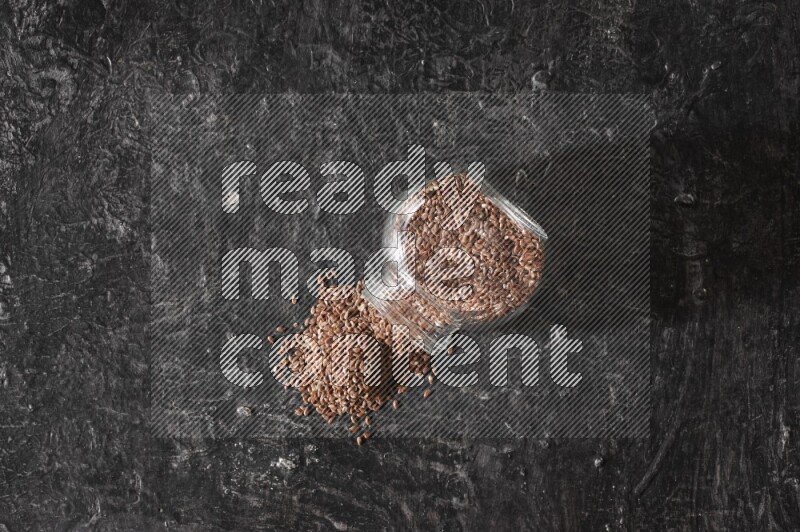A glass spice jar full of flaxseeds flipped and seeds spread out on a textured black flooring