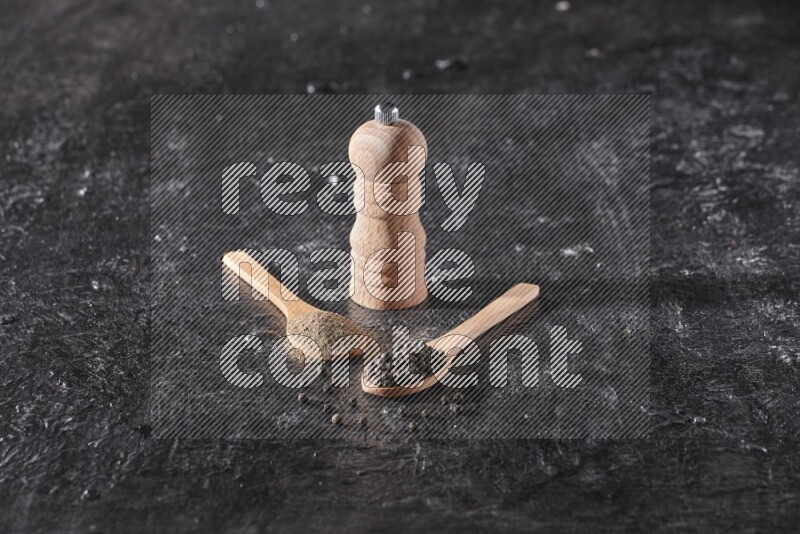 بودرة وحبوب الفلفل الأسود في ملعقتان خشبيتان مع مطحنة خشبية علي خلفية سوداء