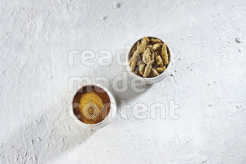2 beige pottery bowl full of turmeric powder and dried turmeric whole fingers on textured white flooring