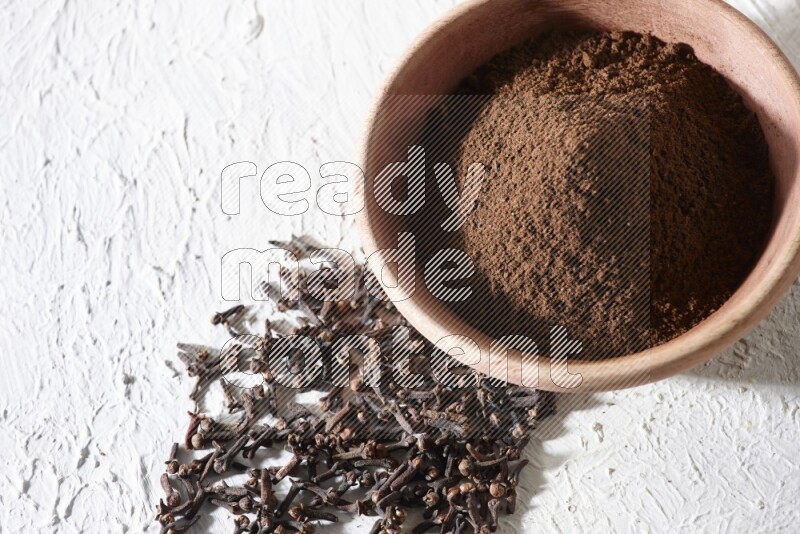 A wooden bowl full of cloves powder with whole cloves beside it on a textured white flooring