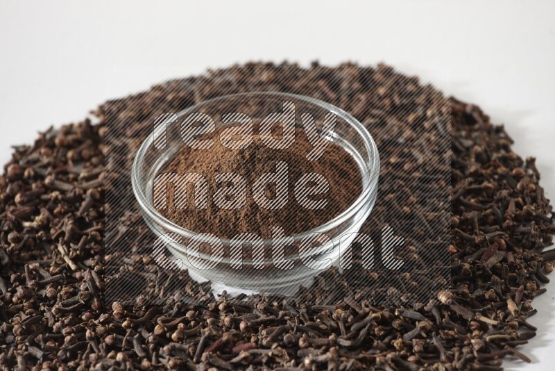 A glass bowl full of cloves powder and cloves grains spread on white flooring