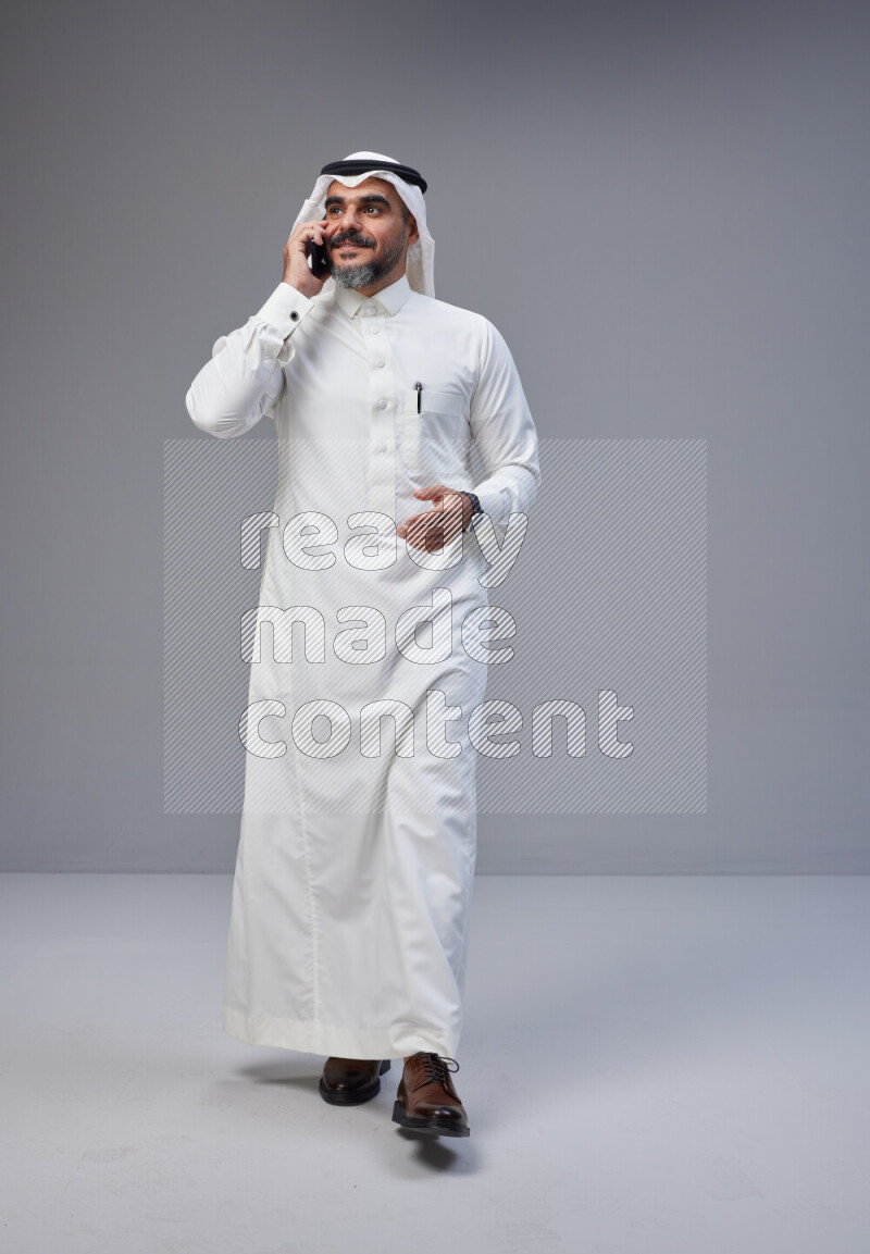 Saudi man Wearing Thob and white Shomag standing talking on phone on Gray background