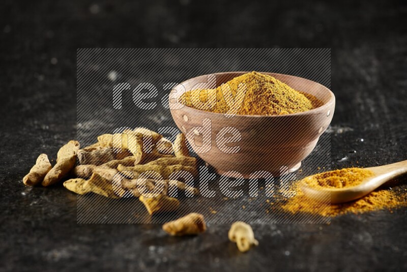 A wooden bowl and a wooden spoon full of turmeric powder with dried turmeric fingers on textured black flooring