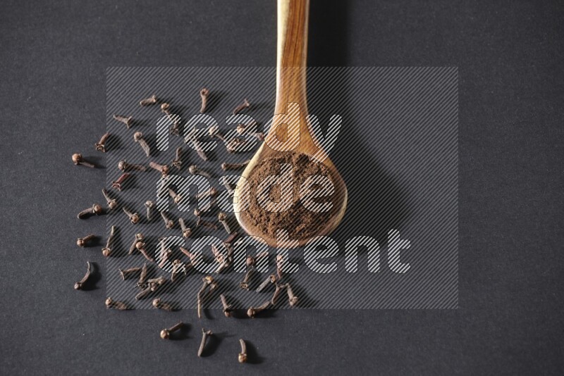 A wooden ladle full of cloves powder and some of whole cloves around it on a black flooring