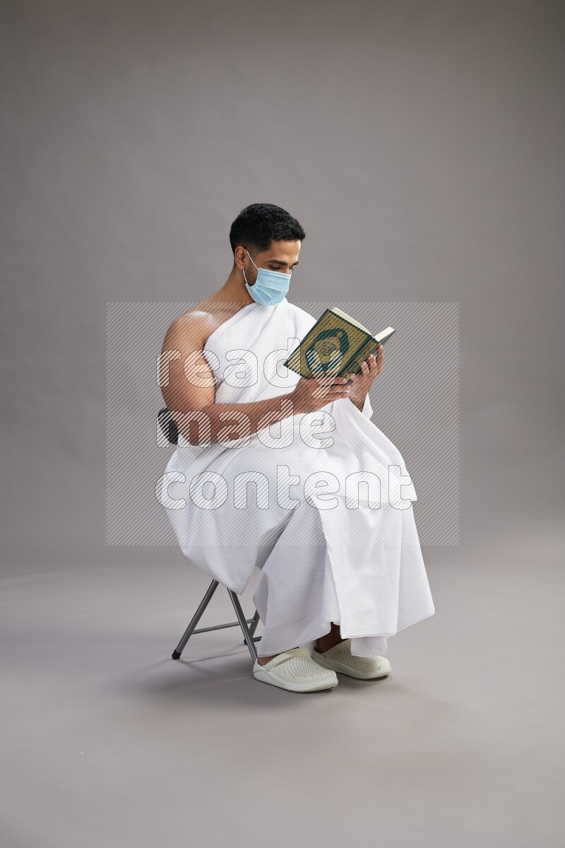 A man wearing Ehram with face mask sitting on chair reading quran on gray background