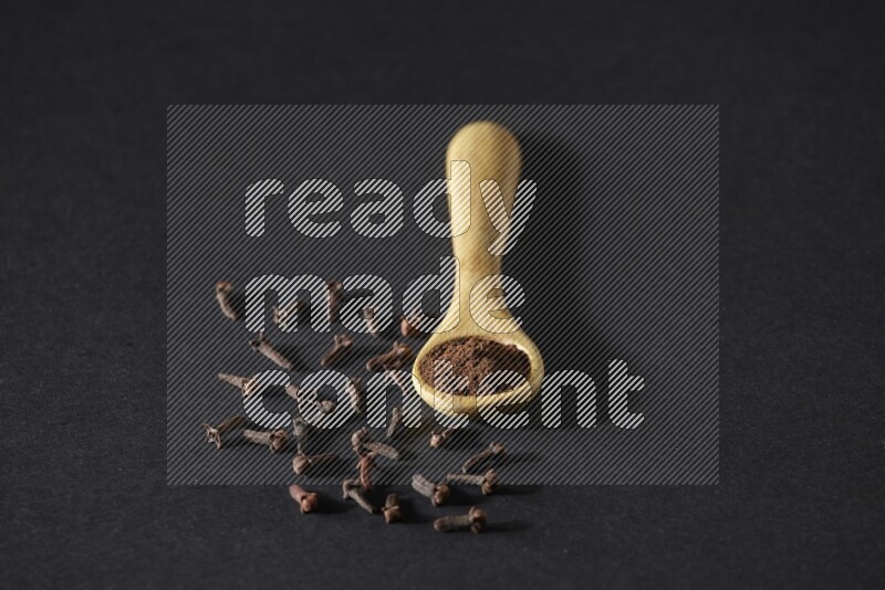 A wooden spoon full of cloves powder with spreaded whole cloves on a black flooring