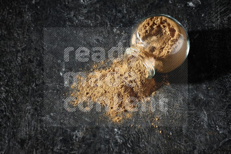 A flipped glass spice jar full of allspice powder and powder spilled out of it on a textured black flooring