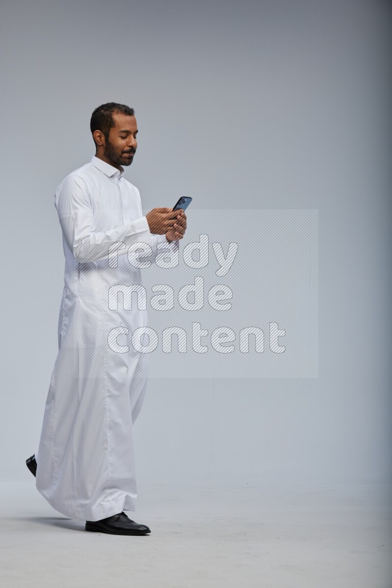 Saudi man wearing Thob standing texting on phone on Gray background