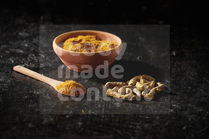 A wooden bowl and a wooden spoon full of turmeric powder with dried turmeric fingers on textured black flooring