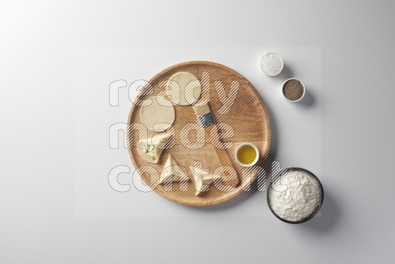 two closed sambosas and one open sambosa filled with cheese while flour, salt, black pepper and oil with oil brush aside in a wooden dish on a white background