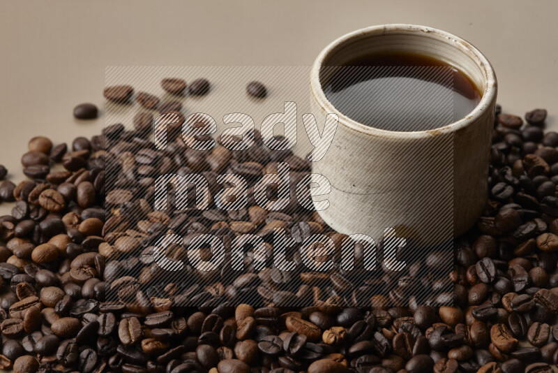 A beige pottery cup of coffee surrounded by roasted coffee beans on beige background