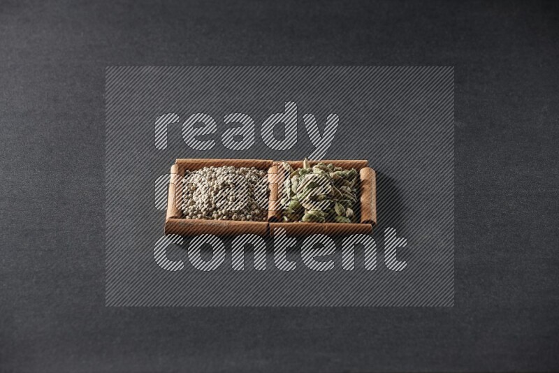 مربعان من أعواد القرفة ممتلئان بحبوب الفلفل الأبيض وحبوب الهيل علي خلفية سوداء