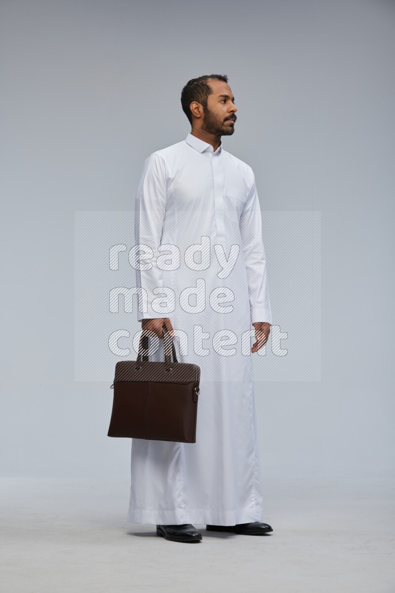 Saudi man wearing thob standing holding suitcase on gray background