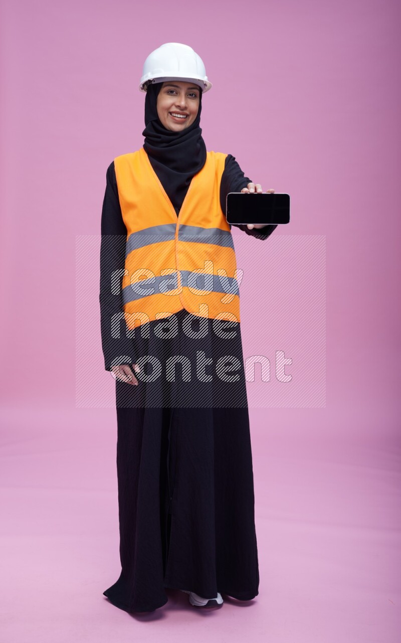 Saudi woman wearing Abaya with engineer vest and helmet standing showing phone to camera on pink background