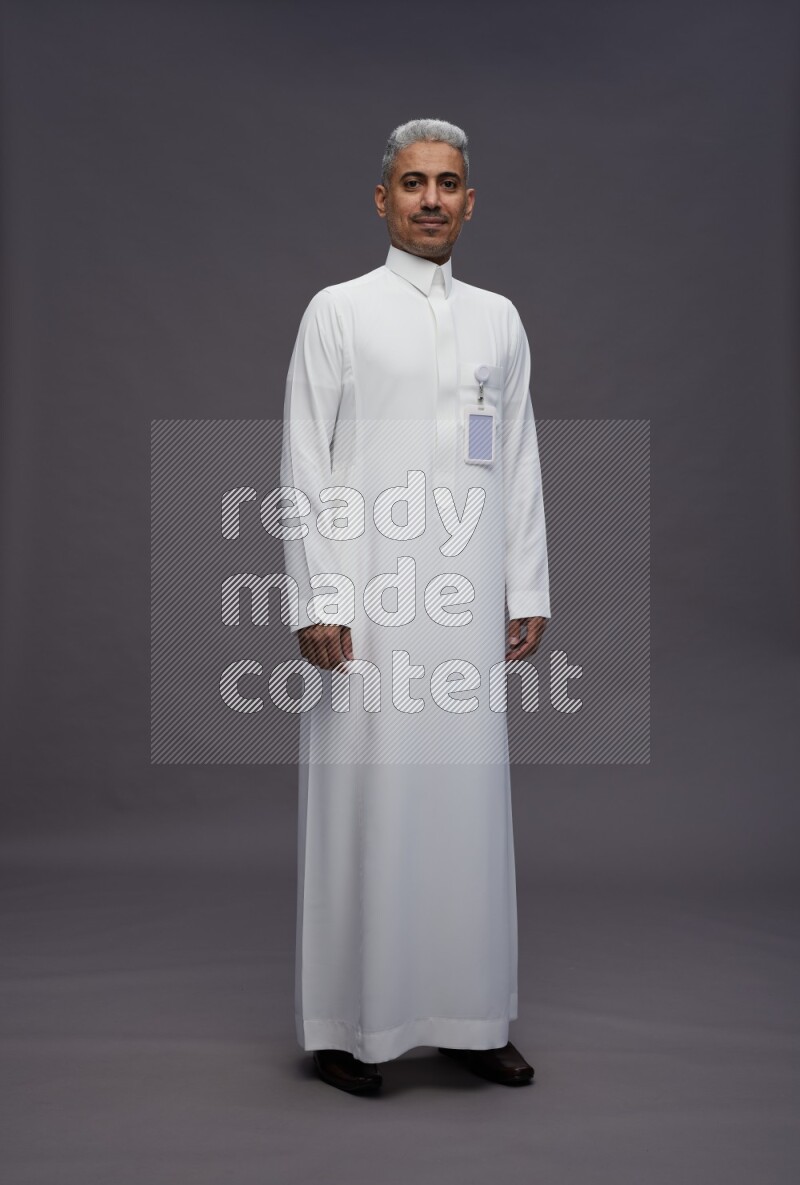 Saudi man wearing thob with pocket employee badge standing interacting with the camera on gray background