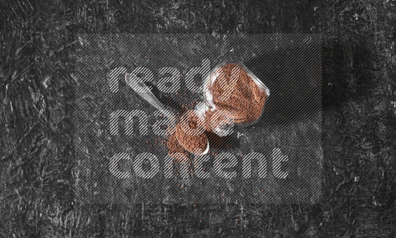A glass spice jar full of garden cress seeds flipped and seeds spread out with a metal spoon full of the seeds on a textured black flooring
