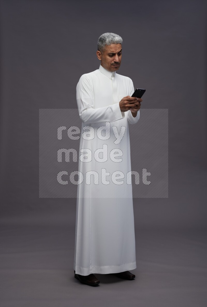 Saudi man wearing thob standing texting on phone on gray background