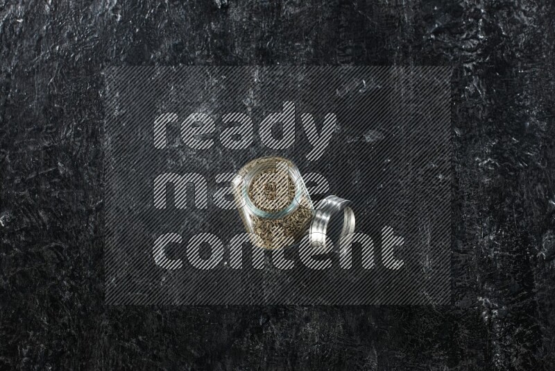 A glass spice jar full of cumin seeds on a textured black flooring