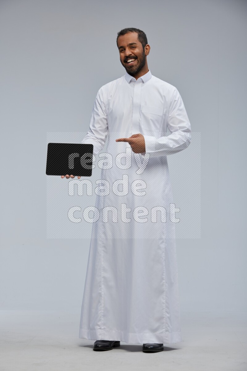 Saudi man wearing Thob standing showing tablet to camera on Gray background