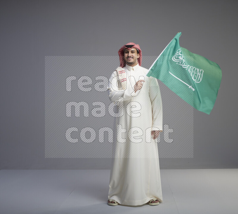 A Saudi man standing wearing thob and red shomag raising big Saudi flag on gray background