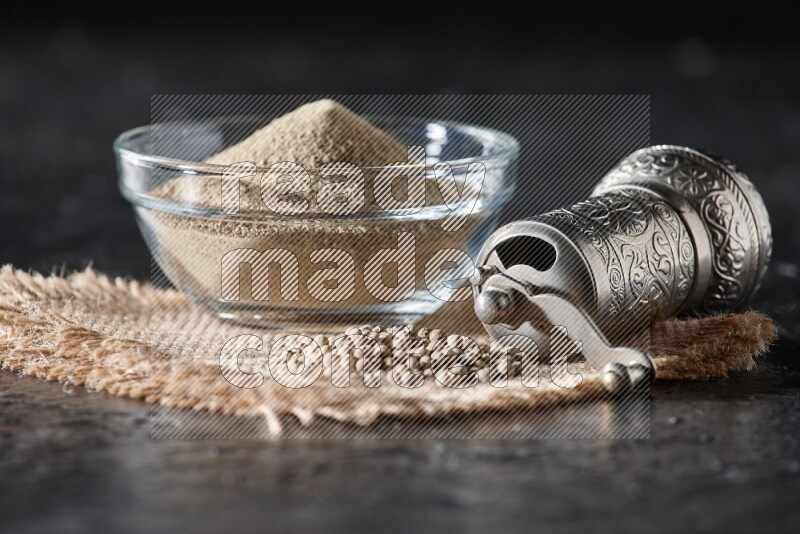 A glass bowl full of white pepper powder with white pepper beads on a burlap piece of fabric and a metal grinder on textured black flooring