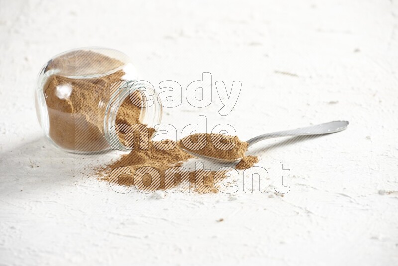Flipped herbs glass jar full of cinnamon powder with a metal spoon full of powder on a textured white background