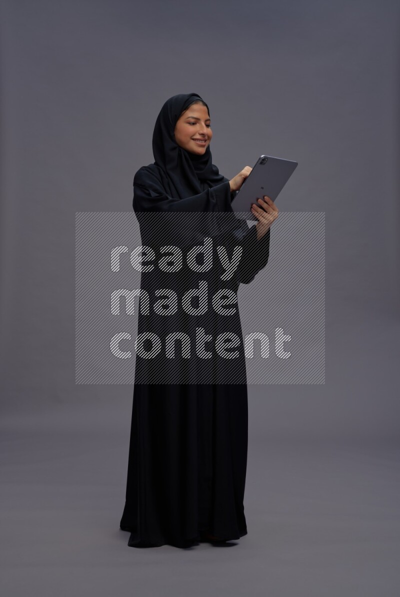 Saudi woman wearing Abaya standing working on tablet on gray background